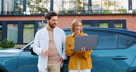 Portrait of woman and man in casual clothes standing in the city near a parked car, discussing future projects and deals using a laptop.
