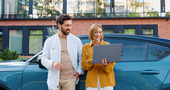 Portrait of woman and man in casual clothes standing in the city near a parked car, discussing future projects and deals using a laptop.