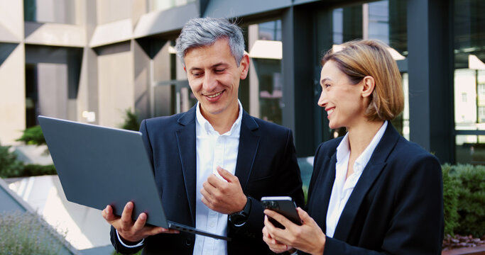 Handheld portrait shot two business partners man and woman in formal suits using laptop and smartphone standing outdoors office building discussing upcoming project and deals. Corporate employees in