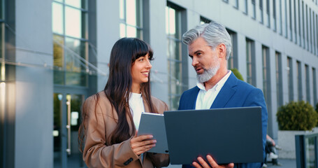 Close-up of mature businessman with laptop and stylish young businesswoman with tablet standing...