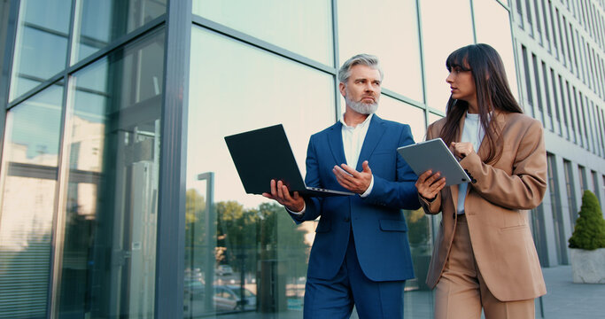 Confident businessman with laptop and elegant businesswoman with tablet walking together down the street toward a modern office building, discussing work and collaborating on a project