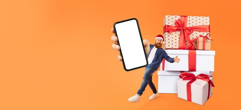 A festive collection showcases a man in a Santa hat holding a smartphone in one hand and standing beside stacked holiday gifts. The orange backdrop adds warmth to the scene.