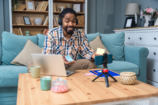 Cheerful creator livestreaming from cozy living room, laptop open, smartphone on tripod, wearing headphones and plaid shirt. Freelancer startup marketing worker video call with client form home office - Powered by Adobe