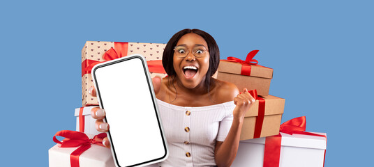 A woman expresses joy while holding a smartphone in front of a stack of wrapped gifts. The backdrop features a bright blue color, enhancing the festive atmosphere of the scene.