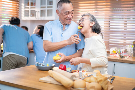 Asian elderly couple enjoying sweet kitchen moments while preparing food. The husband lovingly feeds his wife fruit, reflecting care, companionship, and joyful aging in a warm home.