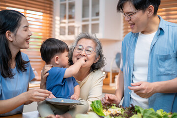 Asian multigenerational family enjoying time together in the kitchen as a toddler lovingly feeds his grandmother. A heartwarming scene showing love, connection, and family bonding across generations.