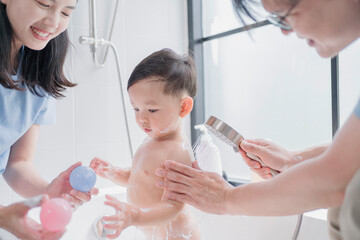 Asian parents giving their toddler a joyful bath, using a handheld shower in a modern bathroom. A happy family moment showcasing love, hygiene, and playful parenting routine.