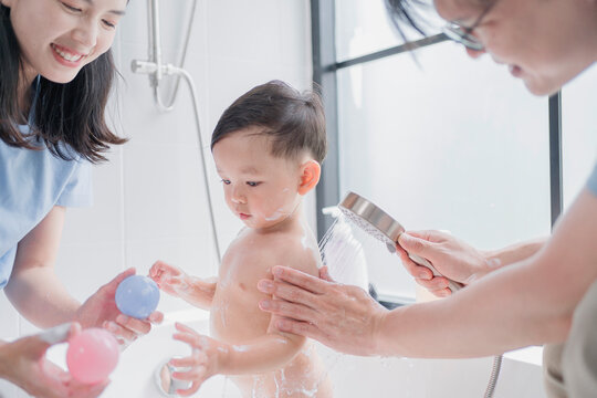 Asian parents giving their toddler a joyful bath, using a handheld shower in a modern bathroom. A happy family moment showcasing love, hygiene, and playful parenting routine. - Powered by Adobe