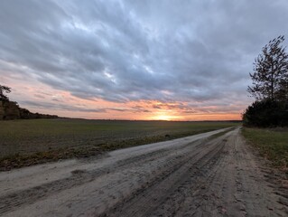 A scenic countryside road bathed in the warm glow of sunset under a dramatic, cloudy sky. The landscape evokes a sense of peace and the beginning of a new day