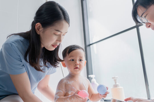 Cute Asian toddler playing with colorful balls in a bubble bath while parents supervise. Adorable moment of baby hygiene, family care, and playful bath time in a modern bathroom.