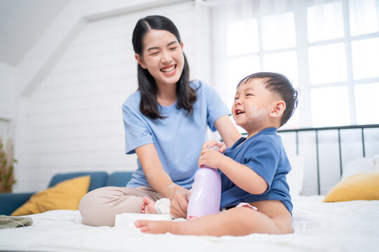 EN: Asian mother smiling while dressing her happy toddler after bath. The baby holds a lotion bottle, capturing a warm family bonding and baby skincare routine in a cozy bedroom. - Powered by Adobe