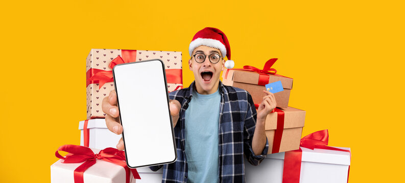 A person wearing a red Santa hat joyfully holds a smartphone and a gift card in front of a colorful stack of presents. The bright yellow background enhances the festive mood.