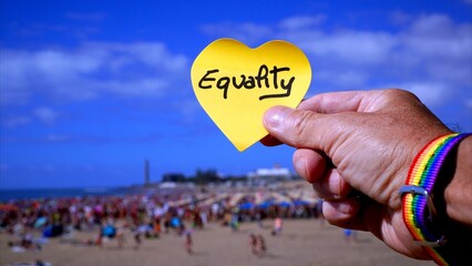 Hand wearing a rainbow bracelet holding a heart-shaped note with the word Equality written on it, symbolizing LGBTQ+ rights and inclusion at a crowded beach event