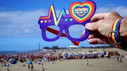 Person wearing rainbow wristband holds novelty pride glasses with heart logo above crowded sunny Maspalomas beach as diverse crowd enjoys summer LGBTQ+ celebration and festival