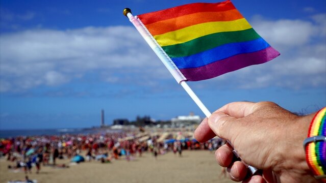 Human hand proudly holding a small rainbow flag with a matching wristband, celebrating on a crowded beach during a gay pride event under a bright blue sky - Powered by Adobe