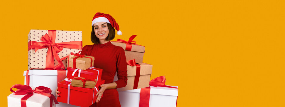 A joyful woman wearing a Santa hat stands amidst various gift boxes wrapped in bright paper with ribbons. She smiles cheerfully, capturing the spirit of the holiday season.