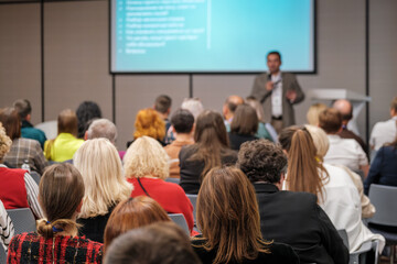 Obraz premium Audience listens to presenter at conference hall with projector screen in background, colorful attendees, business setting, learning atmosphere.