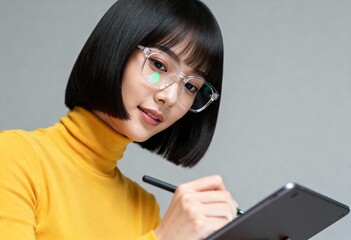 Young Asian woman with glasses drawing on a digital tablet with a stylus. Portrait of a creative female student or designer working on a project against a grey background