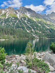 A breathtaking view of a serene lake nestled among towering mountains. The clear water reflects the vibrant greenery and rocky terrain under a bright, blue sky with fluffy clouds.