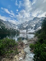 A beautiful lake nestled between tall mountains showcases clear blue water reflecting the cloudy sky. Lush greenery and snow highlight the stunning landscape, creating a peaceful setting.