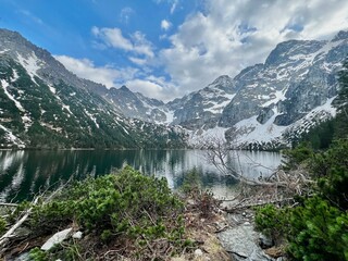 Snow-capped mountains surround a calm lake, reflecting the clouds above. Lush greenery lines the shore, creating a peaceful natural scene in springtime.