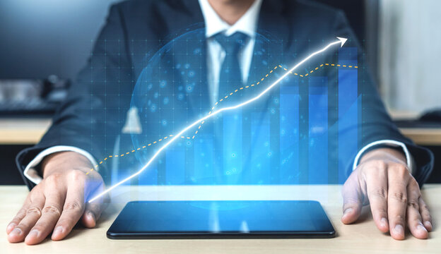 A businessman is analyzing financial data projected from a tablet, showcasing digital graphs that represent positive growth trends and economic forecasting. Impute