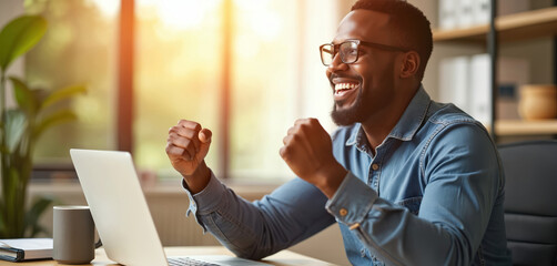 Black man celebrates stock market victory at desk. Smiles intensely reviewing laptop data. Focused investor achieves financial goals through online trading success. Bright workspace reflects