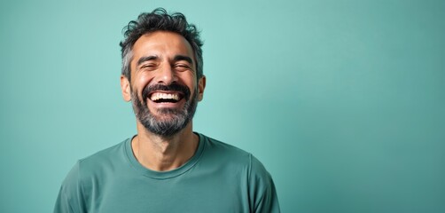 Obraz premium Happy middle eastern man winks broadly, laughing with genuine joy. Dark curly hair with grey streaks, full beard. Wears t-shirt against pastel teal backdrop, conveying warmth, approachability.