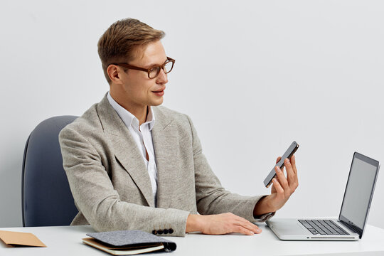 Professional man in formal attire sitting at desk using smartphone with open laptop on a clean, modern office background. Business, work, communication, technology, executive, corporate, male, 30s
