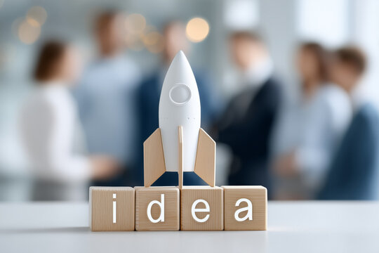 A small rocket sits on wooden blocks that spell the word idea, symbolizing the launch of a new business. In the background, a group of professionals discusses strategies for success and innovation