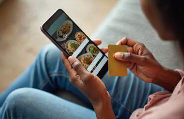 Woman orders food via smartphone app at home. She holds a credit card for online payment. Meal delivery tech service grows in popularity. Mobile shopping concept prospers modernly.