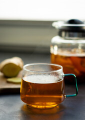 A glas cup of hot tea with fresh ginger and glass tea pot standing on greys kitchen table 
