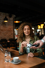 Woman at coffee shop paying with card while sitting at a wooden table with laptop