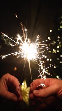 Hands holding glowing fireworks against christmas tree lights bokeh in evening living room. Happy New Year eve video. Atmospheric celebration with burning sparklers
