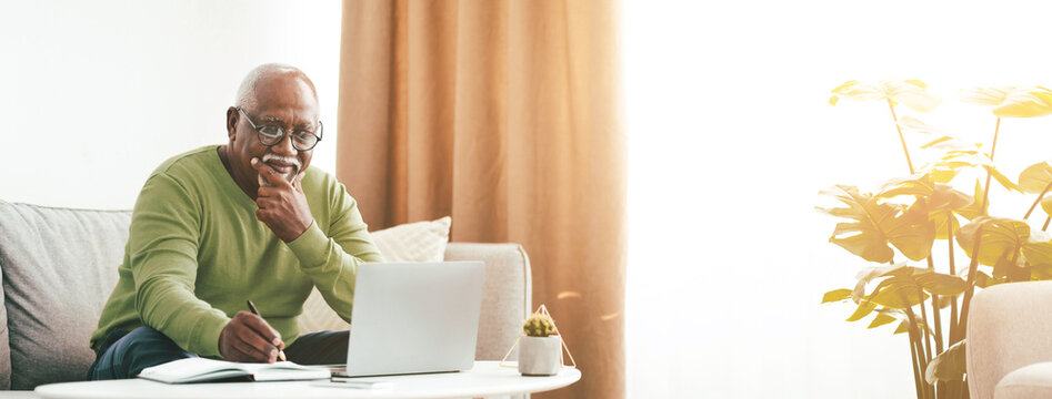 An older man sits on a comfortable couch in bright morning light. He is focused on his laptop, writing notes with a thoughtful expression, surrounded by house plants.