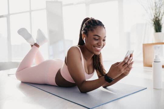 Happy young black woman relaxes on her stomach while using her smartphone to listen to music. She is taking a break from her domestic training workout, showcasing a fit and joyful moment. - Powered by Adobe