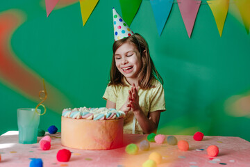 Happy girl wearing party cap sitting at decorated table with cake clapping her hands
