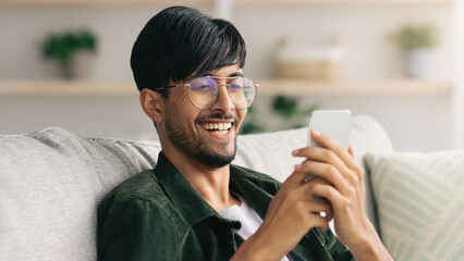 A smiling young man sits on a cozy couch, engaging happily with his smartphone during a relaxed moment at home. Soft natural light fills the room, enhancing the warm atmosphere.