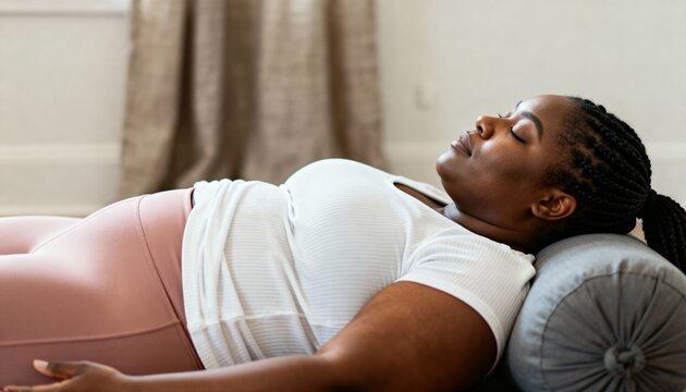 Plus-size Black woman relaxing in Savasana pose on a yoga bolster. Curvy female practicing mindfulness and meditation at home for wellness and self-care