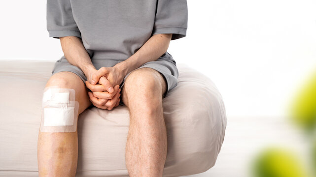 Patient male with bandaged knee sits on bed in light interior. Bruising, medical patch after recent trauma. Concept of injury recovery, first aid, home care, post-surgery healing. Banner. Copy space
