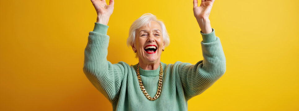 Happy senior woman celebrates with raised hands on yellow background. Cheerful old lady with white hair laughs with joy. Stylish grandmother enjoys success expressing positive emotions, excitement,