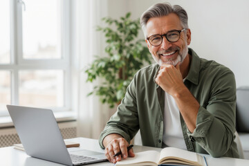 Caucasian man with grey hair and beard wearing glasses and green shirt smiling and looking at camera at desk with laptop and notebook. Remote work and successful business lifestyle, with copy space