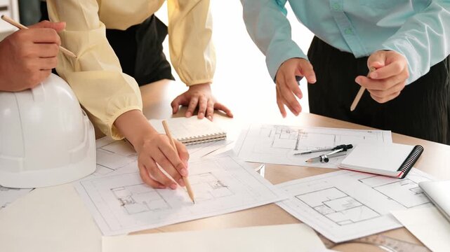 Smart architect and safety engineer discussion about building blueprint on table with architectural document, measurement tools and wooden block scatter around at modern office. Closeup. Delineation.