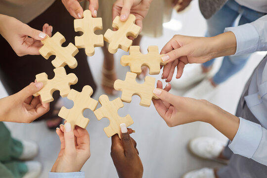 Hands hold puzzle pieces symbolizing teamwork and connection. A diverse group aligns wooden pieces, showing support, unity, and strategy to find a solution. Metaphor of collaboration and unity. - Powered by Adobe