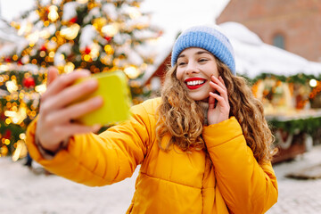 Joyful woman with red lipstick takes selfie at vibrant fair on winter day. Woman in bright jacket...