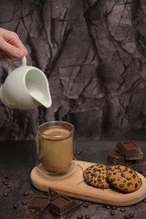 A woman's hand pours milk from a milk jug into a coffee in a glass. Oatmeal cookies on a wooden board. Pieces of broken dark chocolate. Spilled coffee beans. Dark background. Coffee service