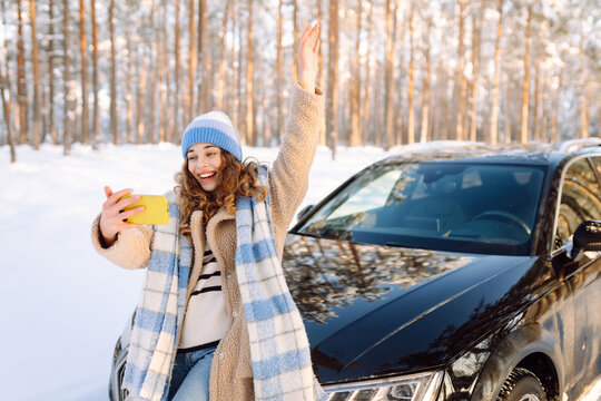 A beautiful woman with phone takes selfie in snowy forest next to car. Young traveler blogs and enjoys sunny winter day. Concepts include blogging, leisure, and road trips. Active lifestyle.