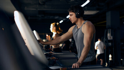 Man with headphones is focused on treadmill in busy gym, working out, panorama. Atmosphere is energetic and lively, typical for afternoon fitness activities