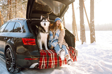 Young woman smiles while sitting in backseat of car with husky on sunny winter day. Woman spends time with pet on road trip, enjoys snowy landscape in forest. Concept: friendship, togetherness, travel © maxbelchenko