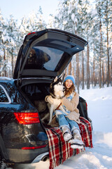 Young woman smiles while sitting in backseat of car with husky on sunny winter day. Woman spends time with pet on road trip, enjoys snowy landscape in forest. Concept: friendship, togetherness, travel